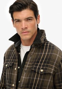 Chaqueta de cuadros marrón hecha de tela texturizada, con acentos amarillos y negros, dos bolsillos en el pecho y un cuello alzado sobre una camisa negra.