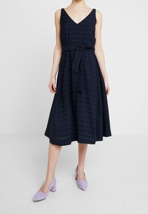 Femme portant une robe midi sans manches bleu marine avec un décolleté en V et une ceinture à la taille, assortie à des chaussures lilas à talons blocs, sur un fond uni.