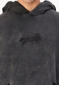 Fekete kapucnis pulóver, megfakult textúrával. Emelt sötét logóval a mellkasán és állítható zsinórral a kapucnin.