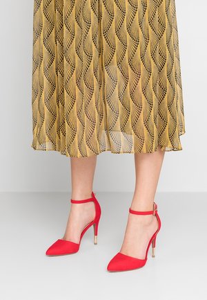 Žena stojící na světlé podlaze před jednobarevným pozadím, má na sobě žlutou vzorovanou midi sukni a červené kotníkové boty na vysokém podpatku s páskem.