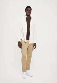 Cămașă ușoară crem, tricou maro, pantaloni bej, adidași albi. Cămașa are închidere cu nasturi în față, iar pantalonii dispun de buzunare laterale și manșete răsucite.