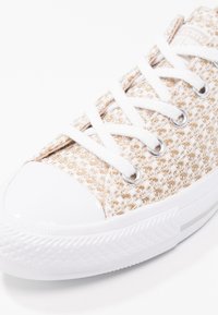 Zapatillas de lona blancas con parte superior texturizada beige, cordones blancos, puntera pulida y ojales plateados. Suela de goma con detalles en patrón.