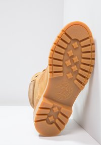 Parte inferior de bota de ante marrón con suela de goma. Características notables incluyen surcos profundos, un logo circular y un tacón ligeramente elevado.