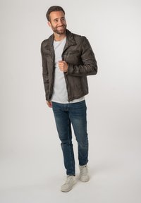 Giubbotto di pelle marrone con tasche con zip, abbinato a una t-shirt bianca e jeans blu. Modello in una posa informale.