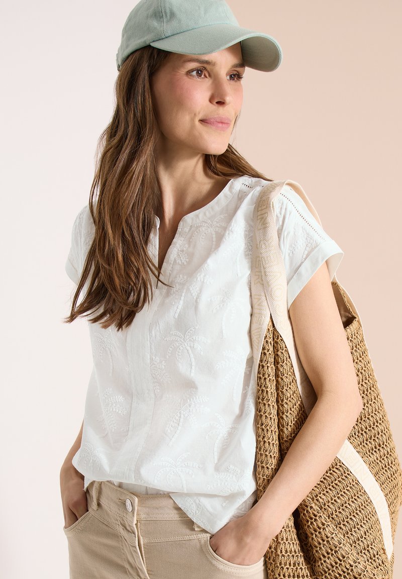 Femme portant une casquette bleu clair, un chemisier blanc brodé à manches courtes, un pantalon beige et portant un sac fourre-tout tissé sur l'épaule.