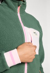 Grønn fleecejakke med rosa glidelås og brystlomme. Har rosa stripete mansjetter og en logo-patch på lommen. Teksturert stoff.