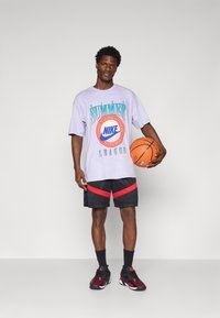 Lyse lilla oversized t-shirt med "Nike Summer League" grafik, sorte shorts med rød stribe detalje og sorte/røde sportssko, mens der holdes en basketball.