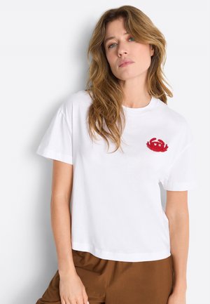 Woman with wavy hair wearing a white T-shirt with a small red crab design on the chest, paired with brown pants, standing against a white wall.