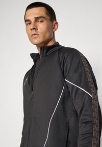 Zwarte sportjack met een hoge kraag, rits aan de voorkant, witte accenten en een patroon langs de mouwen. Gemaakt van glad, lichtgewicht materiaal.