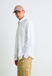 Camisa branca de mangas longas com riscas azuis, com um único bolso no peito, combinada com calças de carga bege com um bolso lateral e o logótipo da Nike. Boné de beisebol bege.