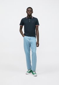 Tmavomodré polo tričko s červenými a bielymi akcentmi na golieri, v kombinácii so svetlomodrými džínsami a zelenými teniskami, stojace proti bielemu pozadiu.