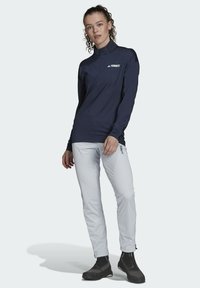 Camisa de manga larga color marino con cuello alto, detalle del logo en blanco. Combinada con pantalones gris claro y botas impermeables negras para uso al aire libre.