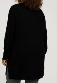 Person mit lockigem Haar, von hinten gesehen, trägt einen langen schwarzen Strickpullover und graue Hosen vor einem einfarbigen Hintergrund.