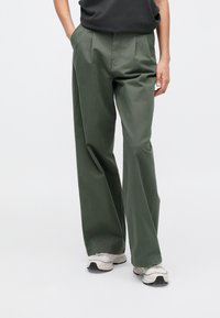 Sötétzöld pamutkeverék bő szárú nadrág, elöl gyűrött, oldalzsebekkel és könnyű anyagból, szürke sportcipővel párosítva.