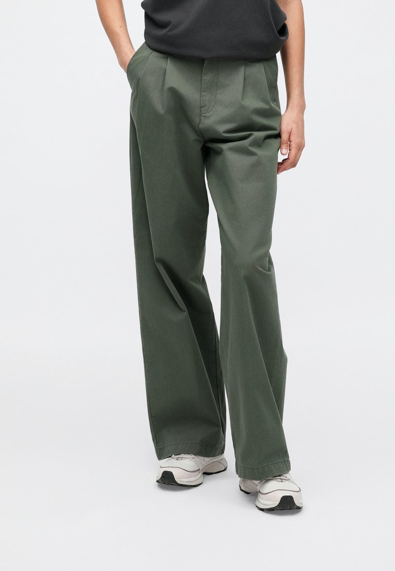 Hose mit weitem Bein aus dunkelgrünem Baumwollmischgewebe, mit vorderen Falten, Seitentaschen und leichtem Stoff, kombiniert mit grauen Sportschuhen.