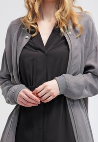 Grå bombarjacka med slät textur, framsida med dragkedja och grå ribbade ärmslut, bärs över en svart klänning med dragsko i midjan.