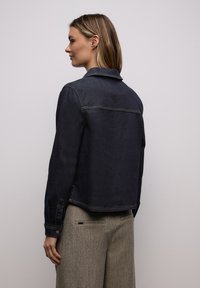 Femme aux cheveux mi-longs portant une veste en jean foncé et un pantalon beige texturé, vue de profil sur un fond uni.