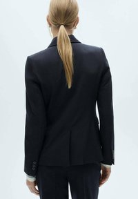 Navy-yhtenäinen bleiseri, joka on valmistettu teksturoidusta kankaasta. Takana on yksi halkio ja hihansuissa kolme nappia. Minimalistinen muotoilu, istuva leikkaus.