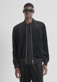 Cazadora bomber de ante negro con cremallera frontal, puños y dobladillo acanalados. Llevada sobre una camiseta negra, combinada con gafas de sol negras.