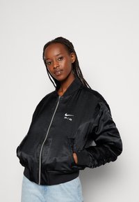 Giubbotto bomber in satin nero con zip frontale, polsini a coste e logo sul petto. Presenta una texture liscia e una vestibilità ampia.