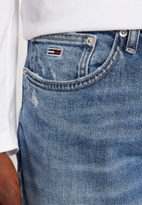Jeans in denim azzurro chiaro con una texture sbiadita, caratterizzati da un piccolo logo ricamato rosso, bianco e blu sulla tasca posteriore e dettagli in metallo.