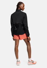 Chaqueta ligera negra con detalles de malla, diseño ajustado y mangas largas, combinada con pantalones cortos coral y zapatillas de running multicolor.