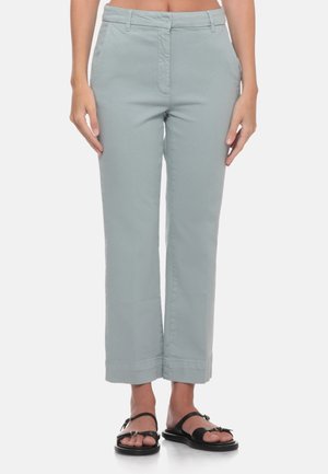 Donna che indossa pantaloni corti grigio chiaro con passanti per cintura e sandali neri aperti in punta, in piedi davanti a uno sfondo bianco.