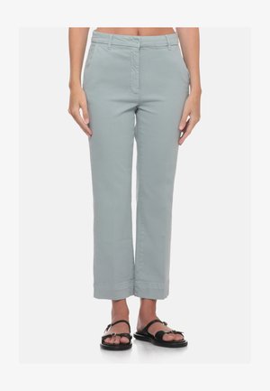 Donna che indossa pantaloni corti grigio chiaro con passanti per cintura e sandali neri aperti in punta, in piedi davanti a uno sfondo bianco.