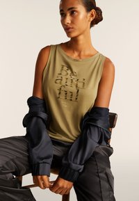 Top sin mangas verde oliva con la palabra "Beautiful" en letras estilizadas, combinado con una chaqueta bomber azul marino y pantalones negros. Tela suave, corte relajado.