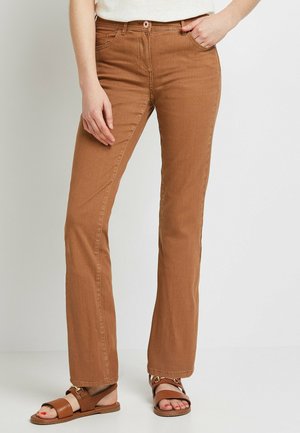 Kobieta ubrana w brązowe spodnie z wysokim stanem i prostymi nogawkami z przednimi kieszeniami, w połączeniu z brązowymi sandałami i białą bluzką.