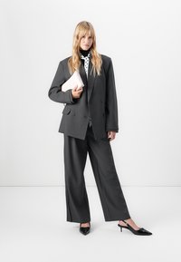 Frau mit langen blonden Haaren, die einen übergroßen dunkelgrauen Anzug und schwarze spitze High Heels trägt, hält eine blassrosa Clutch vor einem weißen Hintergrund.
