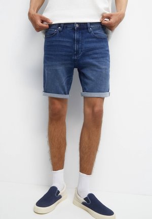 Dunkelblaue Denim-Shorts mit umgeschlagenem Saum und Fünf-Taschen-Design, getragen mit weißen Knöchel-Socken und marineblauen Slippern.