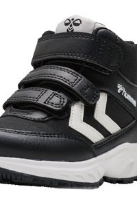 Zapatilla alta negra con tiras de Velcro, parte superior de cuero texturizado, acentos en forma de cheurón blanca y una suela de goma con patrones de tracción.