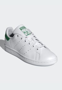 Hvit lær sneakers med grønne detaljer, med snøring, perforerte detaljer på siden og en logo-patch på tungen.