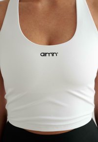 Fehér sport crop top széles nyakkivágással és a középen elhelyezkedő fekete "aim'n" logóval. Sima textúrájú, testhezálló dizájn, fekete alsóneművel párosítva.