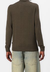 Pull vert olive avec col et poignets côtelés, présentant une texture lisse. Associé à un jean en denim clair, affichant une coupe décontractée.