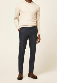 Béžový vlněný svetr s kulatým výstřihem, spárovaný s tmavě modrými kalhotami na míru. Hnědé chelsea boty doplňují celý vzhled.