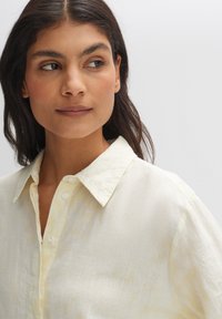 Light yellow button-up shirt with a textured fabric, featuring a pointed collar and a relaxed fit. Subtle vertical stripes are visible.