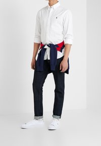 Biała koszula na guziki, niebiesko-czerwony sweter związany w supeł wokół talii, ciemne dżinsy, białe tenisówki; codzienny strój z klasycznymi elementami.