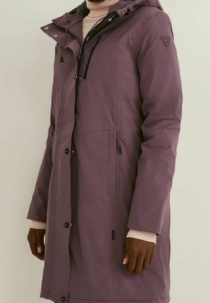 Personne portant un long manteau d'hiver mauve avec une capuche, des boutons sur le devant, des poches latérales zippées, et des manches de pull beige visibles aux poignets.