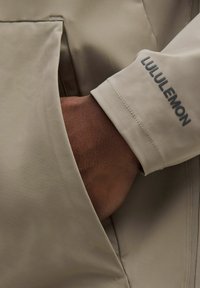 Beige Jacke mit strukturiertem Finish und sichtbarem Taschen, mit einem Grauen Aufnäher am Bündchen, auf dem "LULULEMON" steht. Nahaufnahme der Handinteraktion.