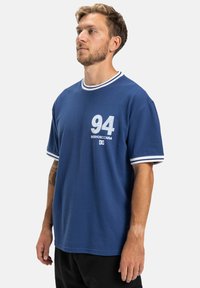 Modrá polokošeľa s krátkym rukávom a bielym žebrovaným golierom a manžetami. Na prednej strane je potlač "94" v svetlosivej farbe. Tkanina vyzerá hladko a mäkko.