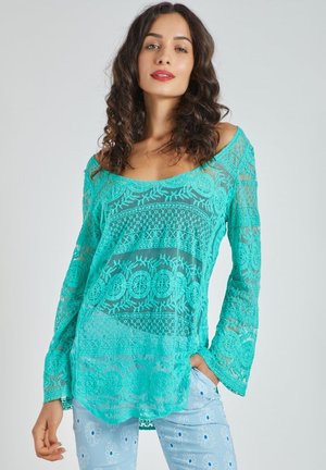 Top de encaje turquesa con patrones florales y geométricos, escote amplio, mangas largas acampanadas, textura translúcida, combinado con pantalones estampados en azul claro.