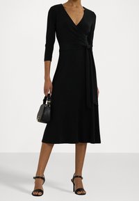 Černé zavinovací šaty s V-výstřihem, tříčtvrtečními rukávy a detailem uvázání v pase. Doplněné malou černou kabelkou a sandály na řemínek.