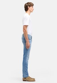 Jasnoniebieskie dżinsy o wąskim kroju, z klasycznymi kieszeniami i subtelnym przeszyciem z tyłu. Noszone z prostą białą koszulką i beżowymi butami.