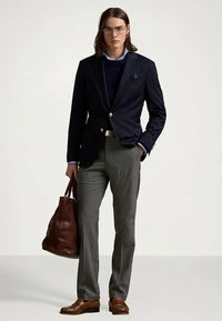 Ung man i glasögon, iklädd navyblå blazer, ljusblå skjorta, grå byxor och bruna läderskor, hållande en stor brun läderväska.