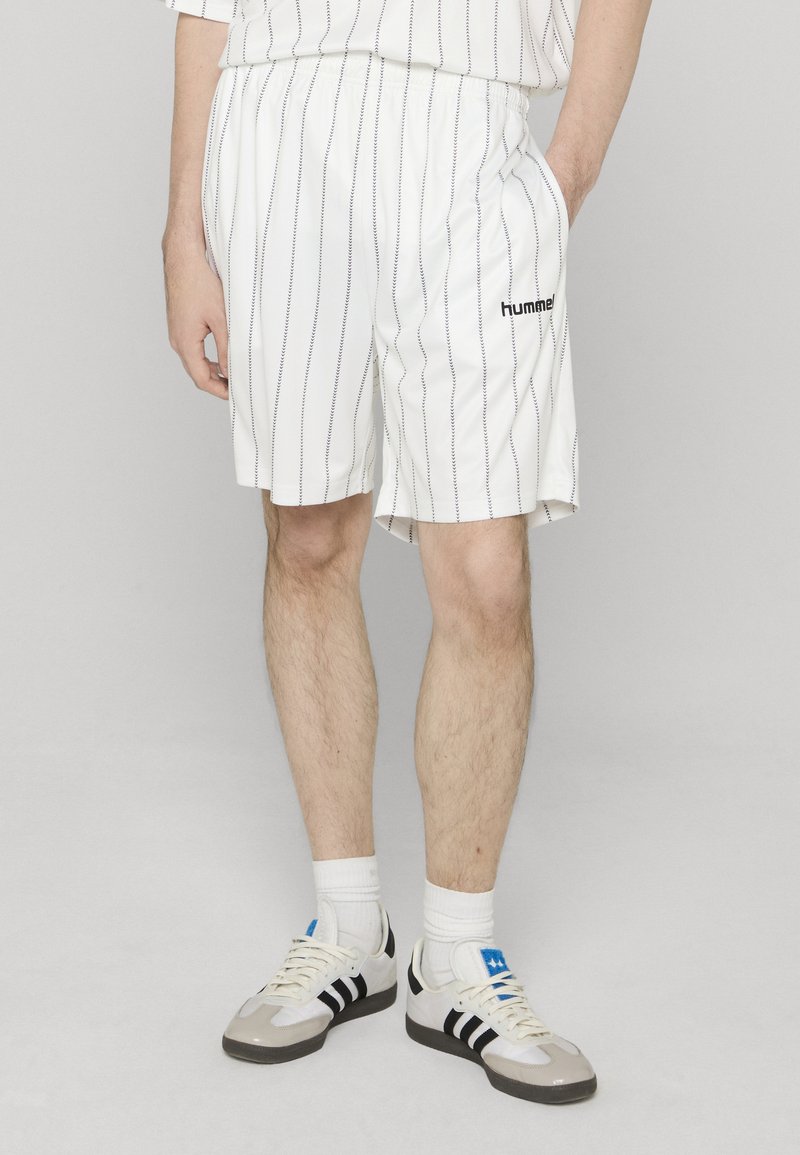 Shorts de sport blancs avec des fines rayures bleues, taille élastique, poches latérales, et un logo "hummel" noir près de l'ourlet. Portés avec des baskets.
