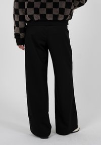 Person, die schwarze Hosen mit weitem Bein und einen braun-schwarz karierten Pullover trägt, stehend vor einem schlichten hellen Hintergrund.