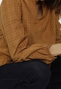 Blusa marrone a quadri con vestibilità ampia, maniche lunghe arricciate ai polsini e scollatura arrotondata. Indossata sopra pantaloni in denim scuro.