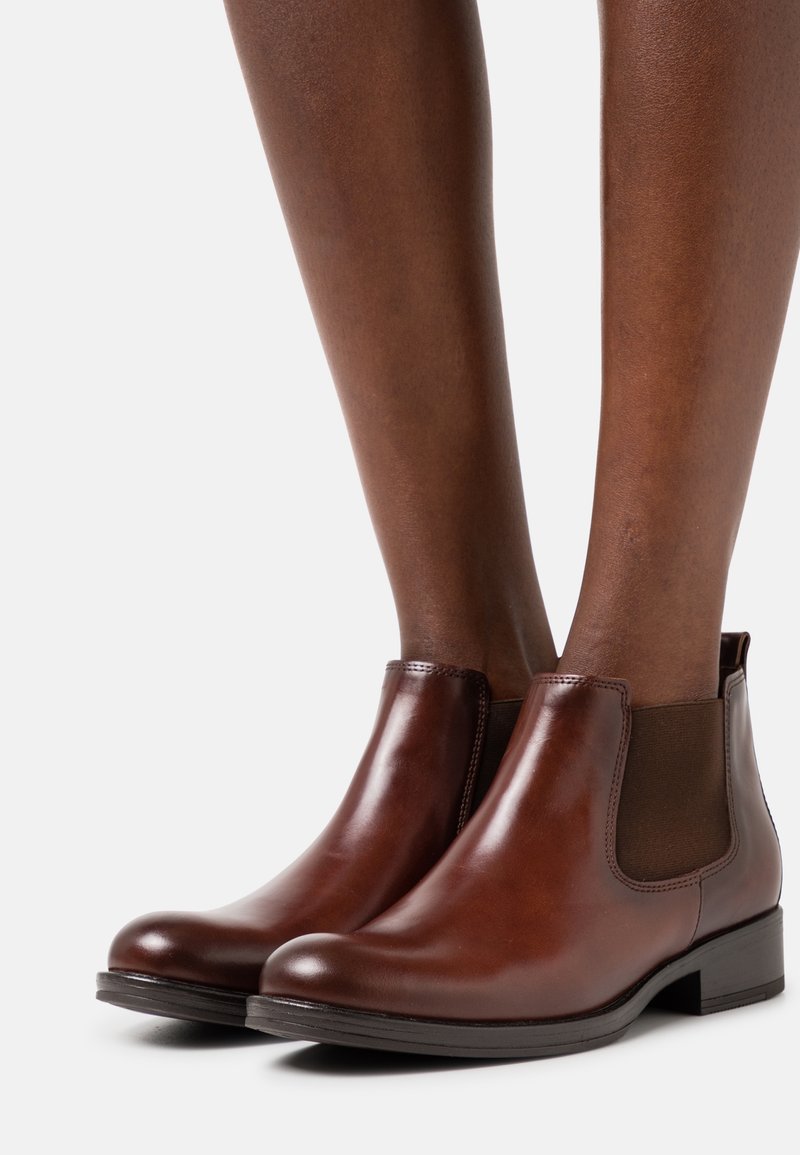 Bottes Chelsea en cuir marron avec panneaux latéraux élastiques portées sur des jambes à la peau foncée sur un fond blanc.